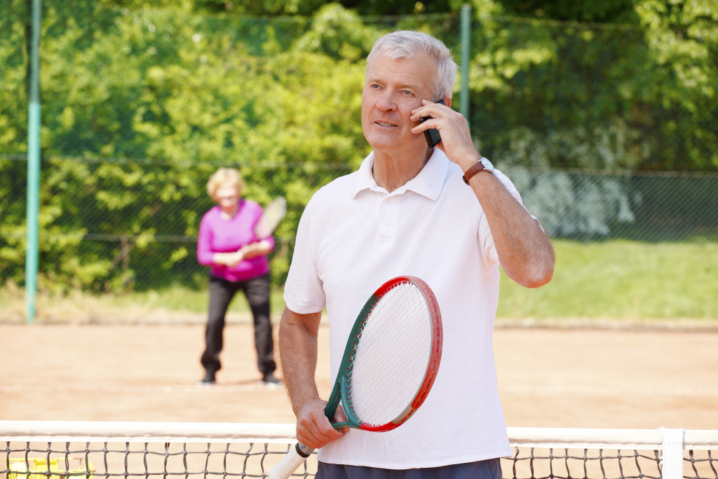 an elderly man talking to the phone with a tennis racket in hand