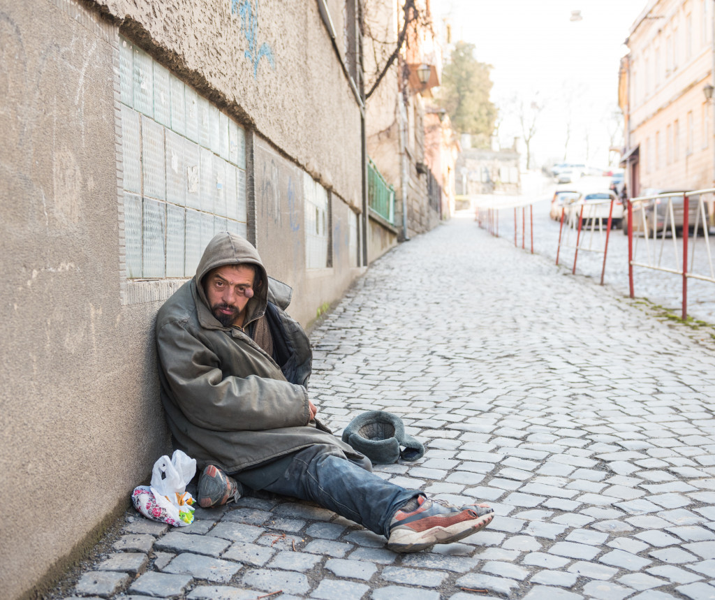 A homeless man sitting on the sidewalk with his possession beside him.